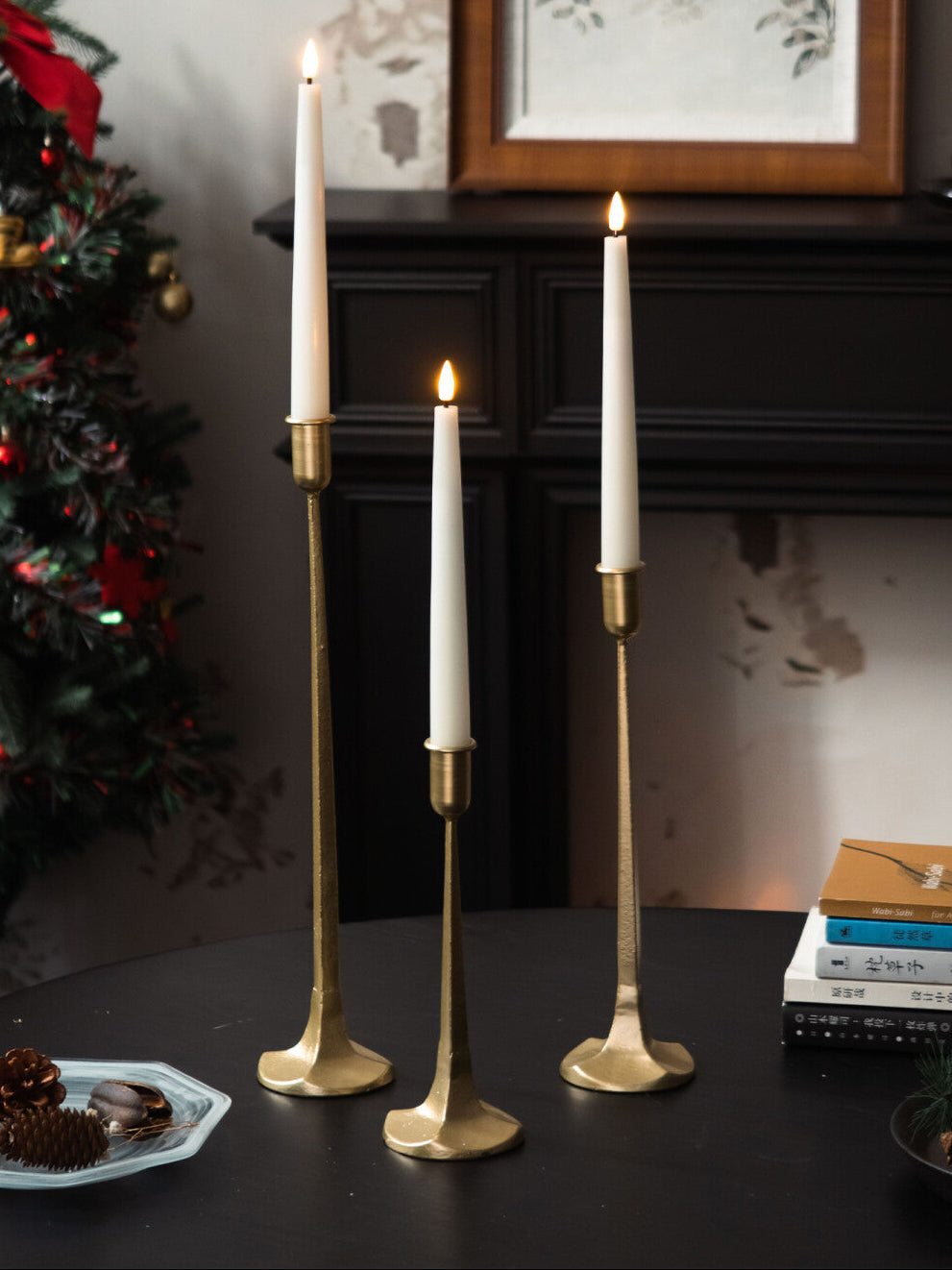 Three gold candle holders with white candles on a dark surface, with a Christmas tree and framed artwork in the background.