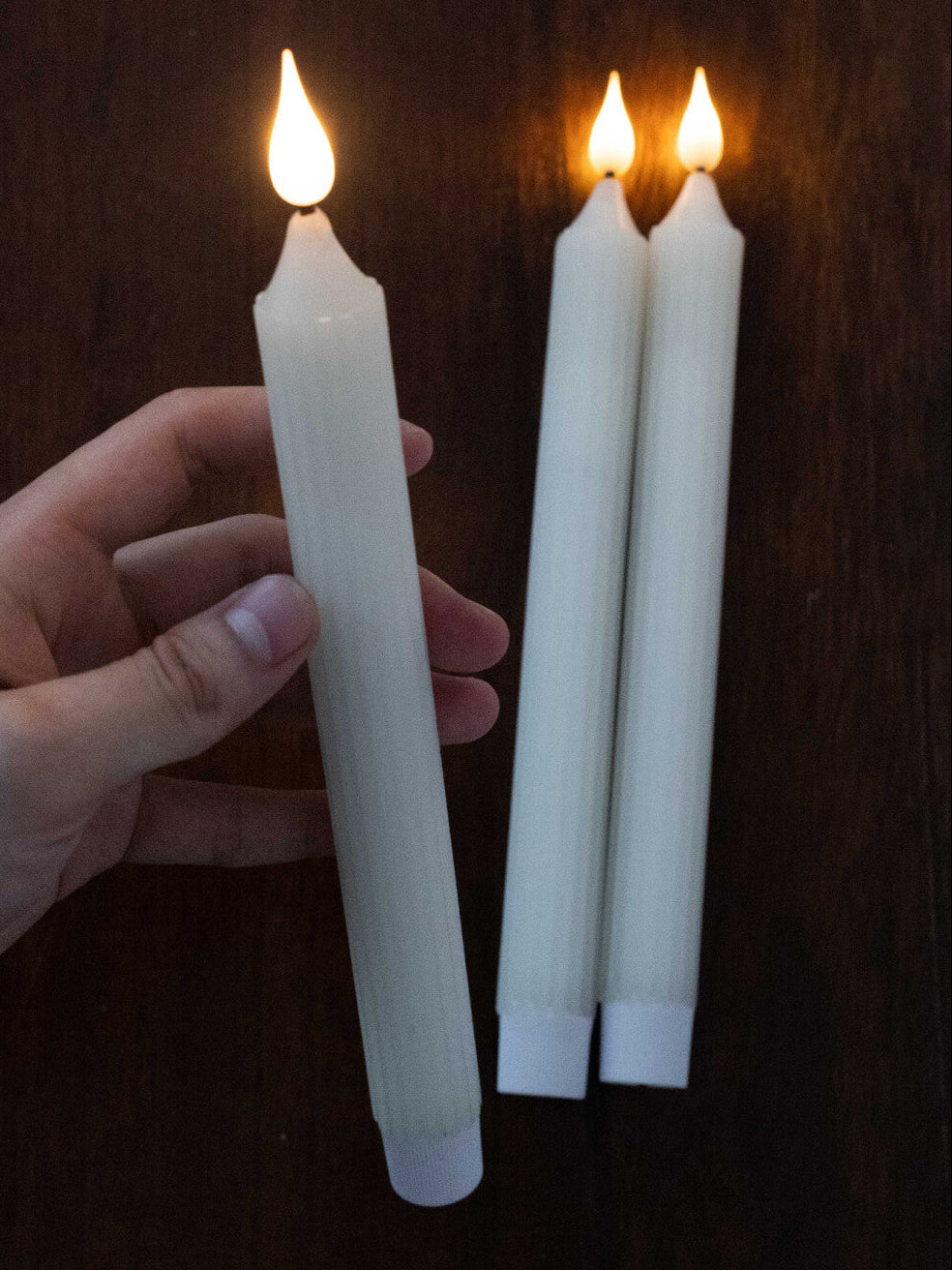 Hand holding a lit white candle with three more candles in the background on a dark surface.