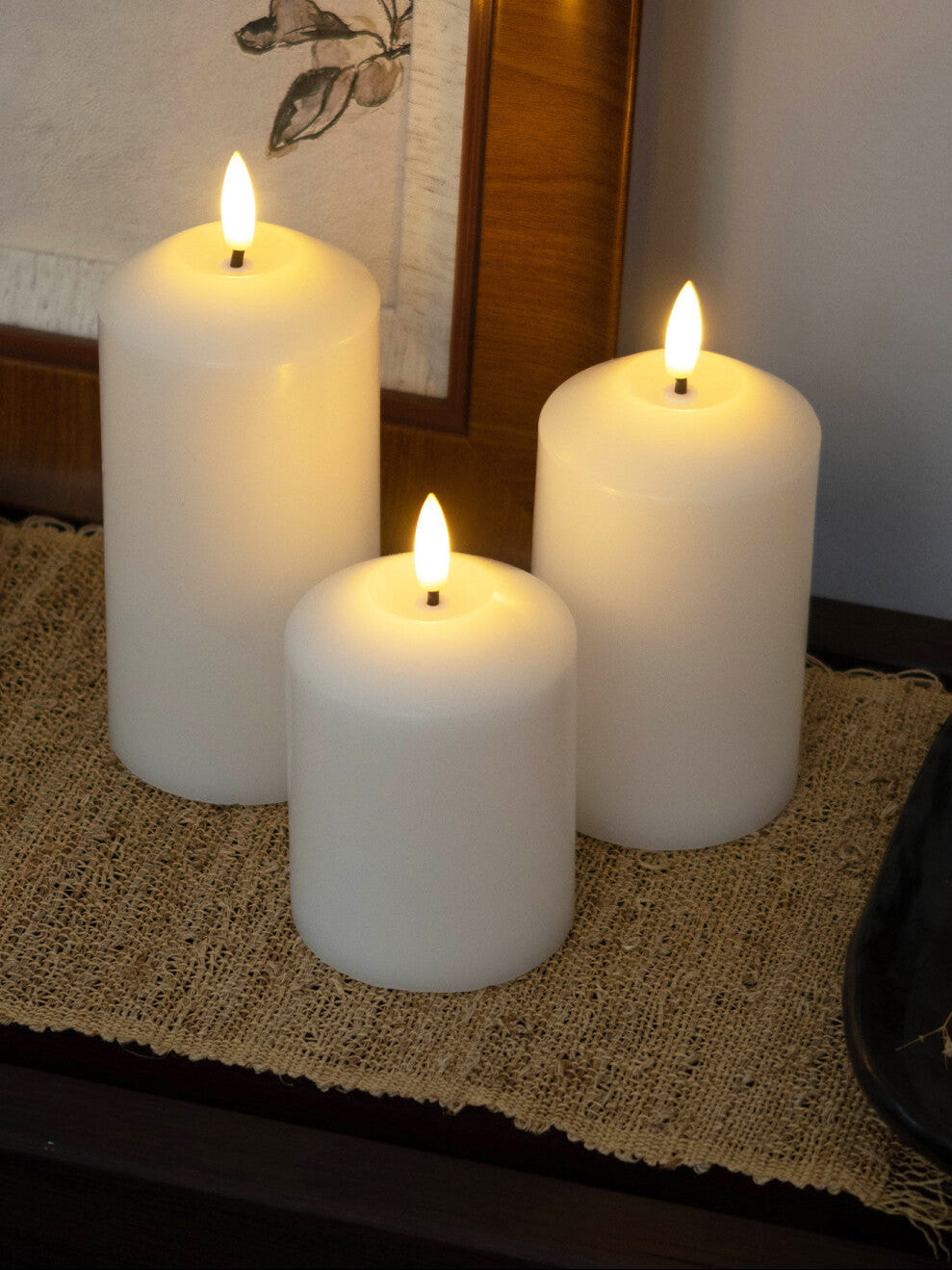 Three white candles with flame on a textured surface, possibly a table or mat.