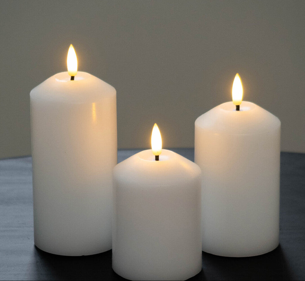 Three white candles of different sizes with a flame on a dark surface and gray background.