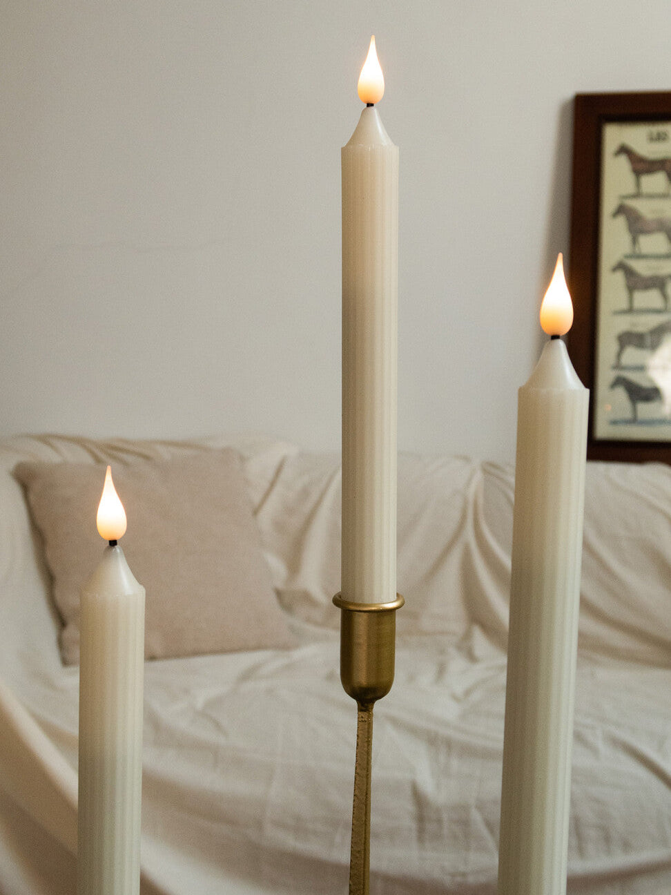 Three lit candles in front of a white sofa with a decorative frame on the wall.