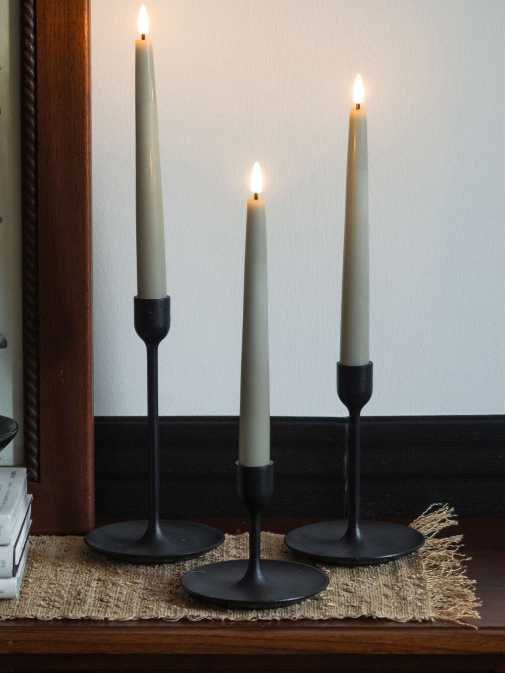 Three lit candles in black holders on a wooden surface with a white wall background.