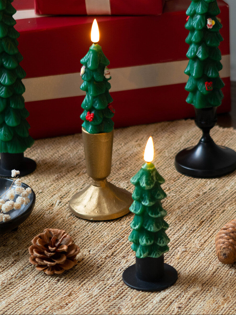 Decorative Christmas tree-shaped candles on a textured surface with pine cones and a gift box in the background.